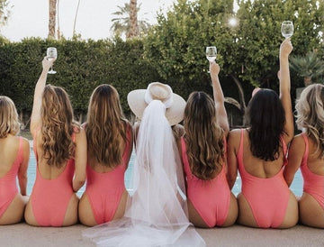 Group of women sitting poolside with their backs to the camera, all wearing matching pink swimsuits. The bride, in the center, is wearing a white sunhat and veil. They are raising glasses in celebration, with palm trees and greenery in the background.