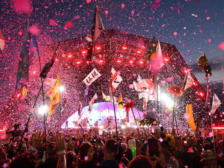 Crowd at Glastonbury Festival cheering under confetti with flags waving and stage lights glowing at sunset.