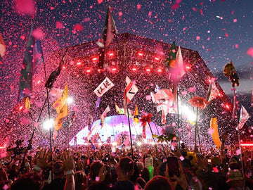Crowd at Glastonbury Festival cheering under confetti with flags waving and stage lights glowing at sunset.