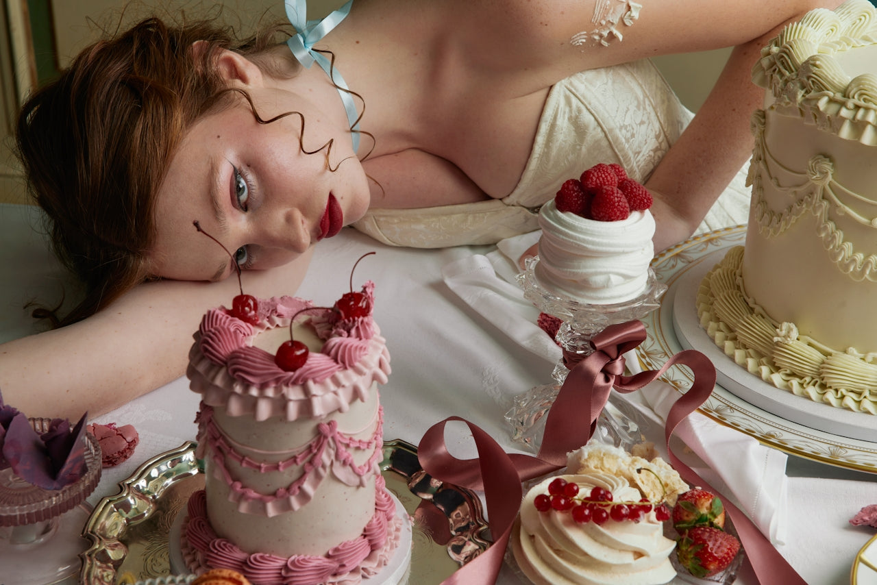 Woman in a cream dress lying on a table surrounded by ornate vintage cakes and desserts, styled in a dreamy, decadent setting.