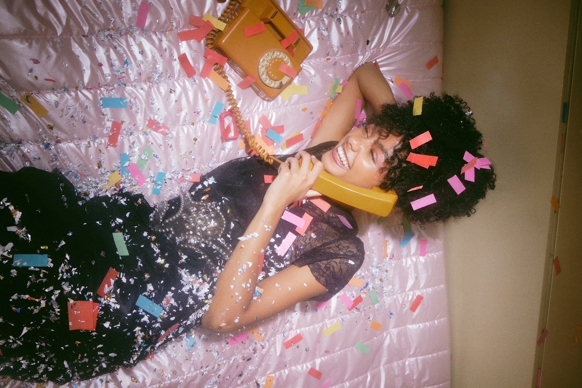 Woman lying on a satin pink bedspread, smiling while talking on a vintage yellow rotary phone, surrounded by colorful confetti. She is wearing a sparkly black dress and has confetti in her curly hair, creating a joyful, party-like atmosphere.