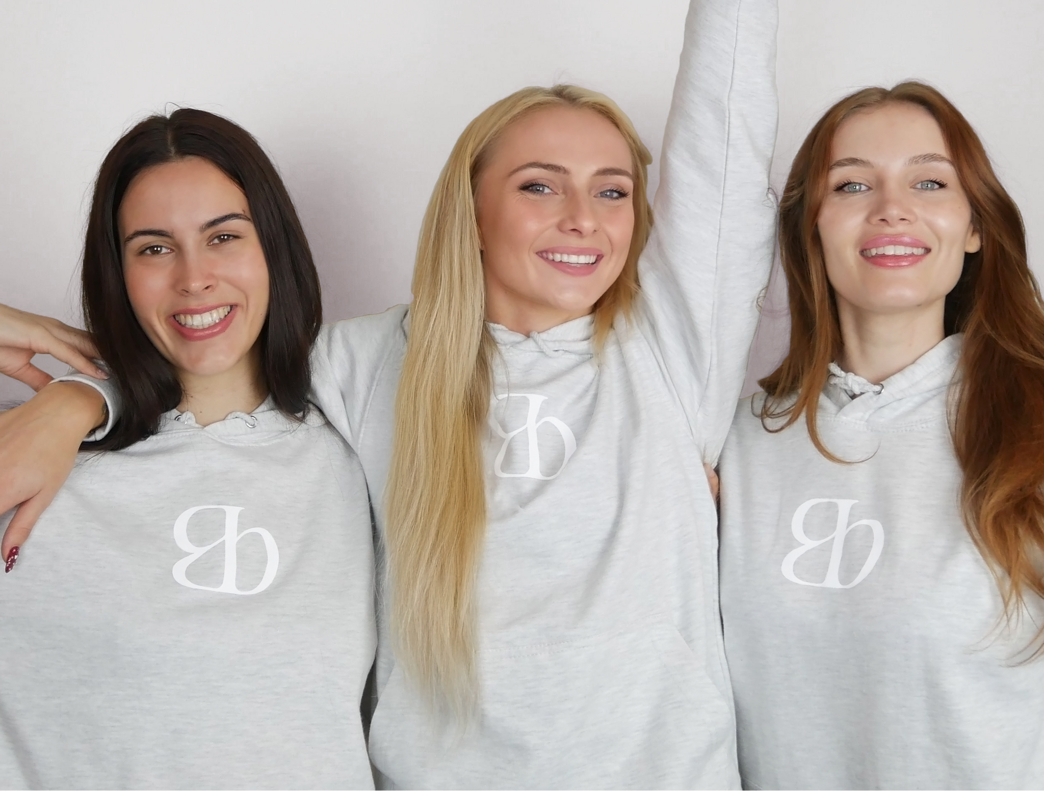 Three smiling women wearing wellness hoodies, celebrating together and holding a smoothie. A joyful moment of community and vitality.