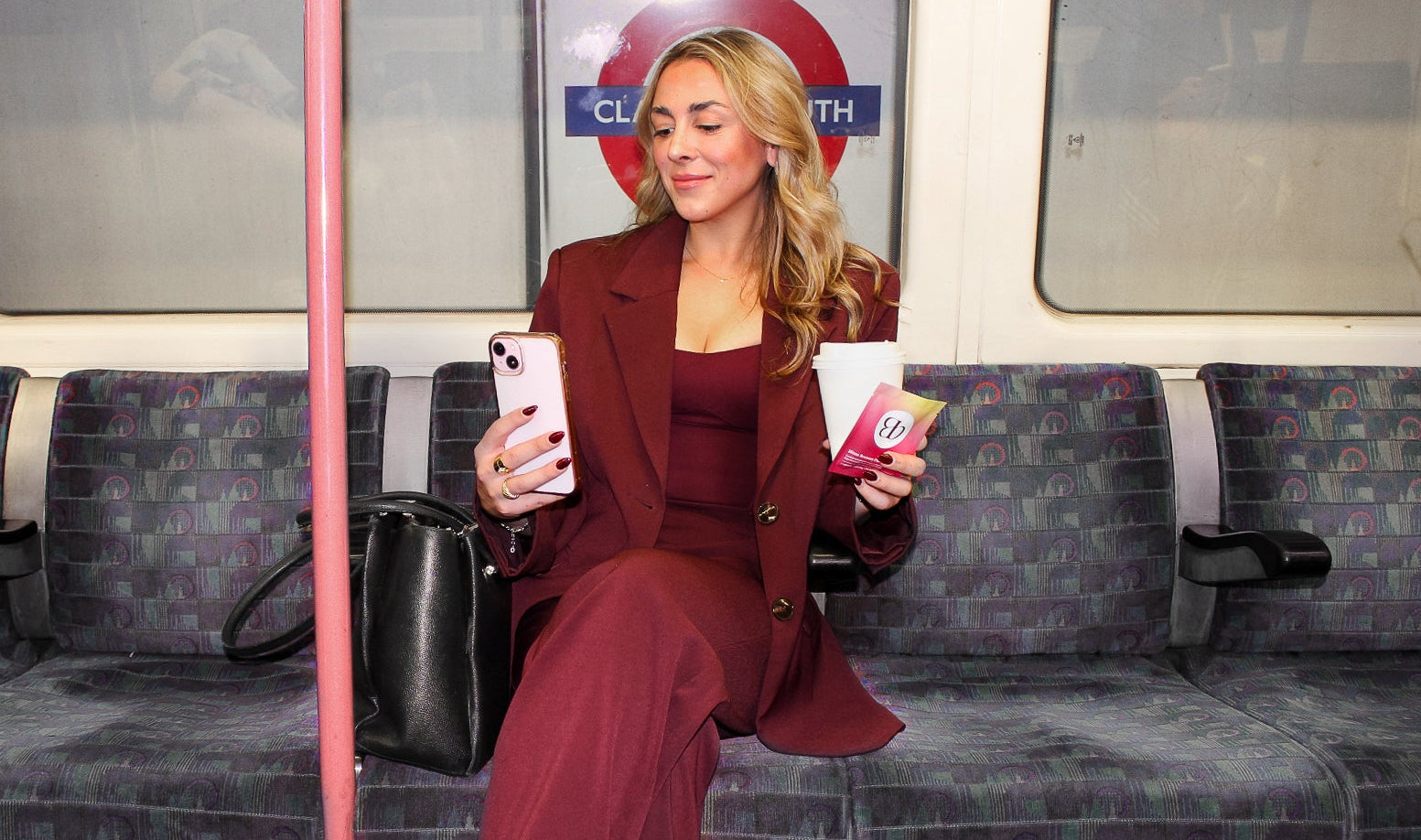 Woman in a burgundy suit on the London Tube holding coffee and a Blōma sachet after work, staying fresh for next-day productivity.slider_item_VVWPnc